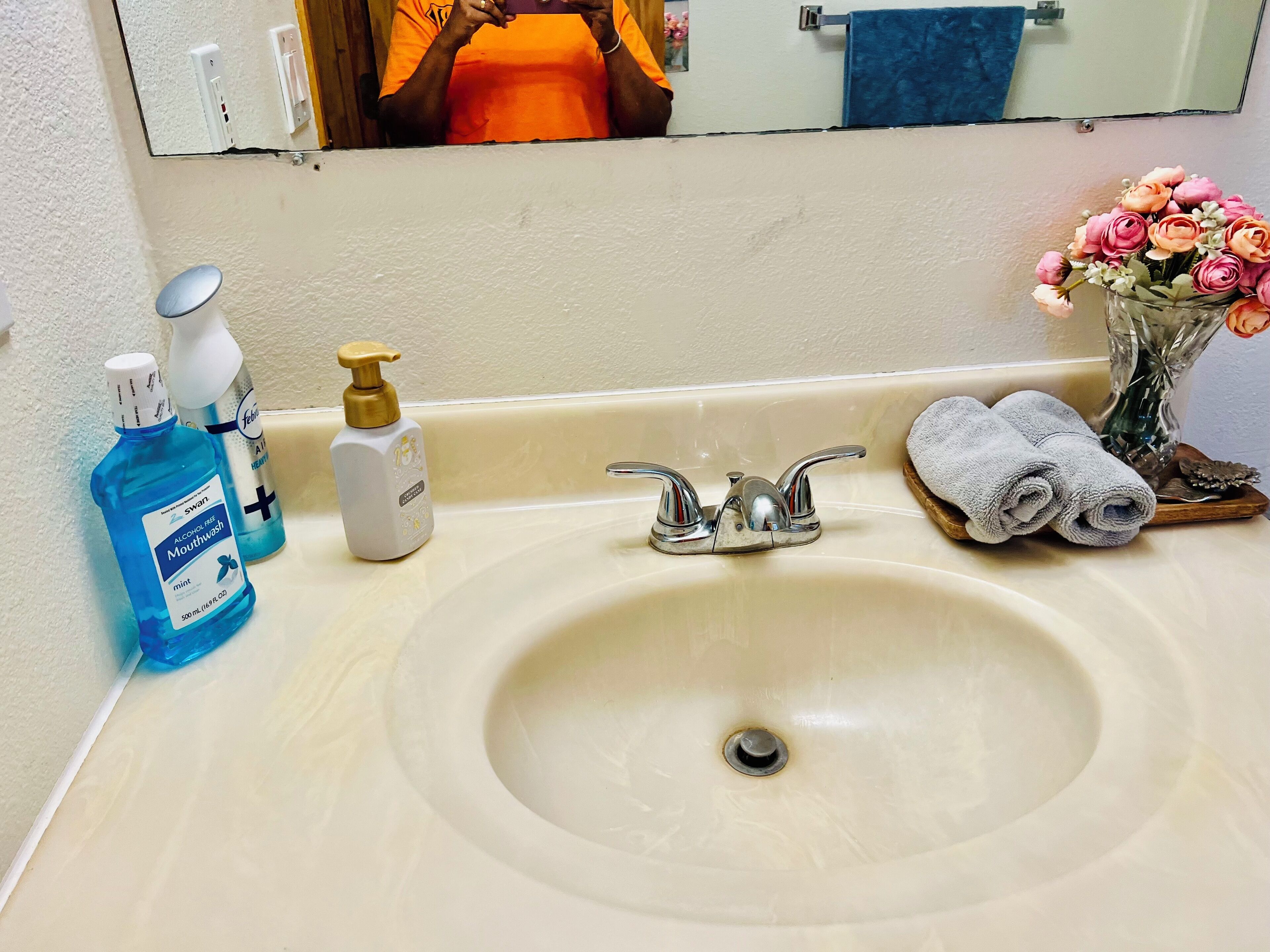Ein Waschtisch mit Spiegel, Mundspülung, einer Shampooflasche und einer Vase mit Blumen.