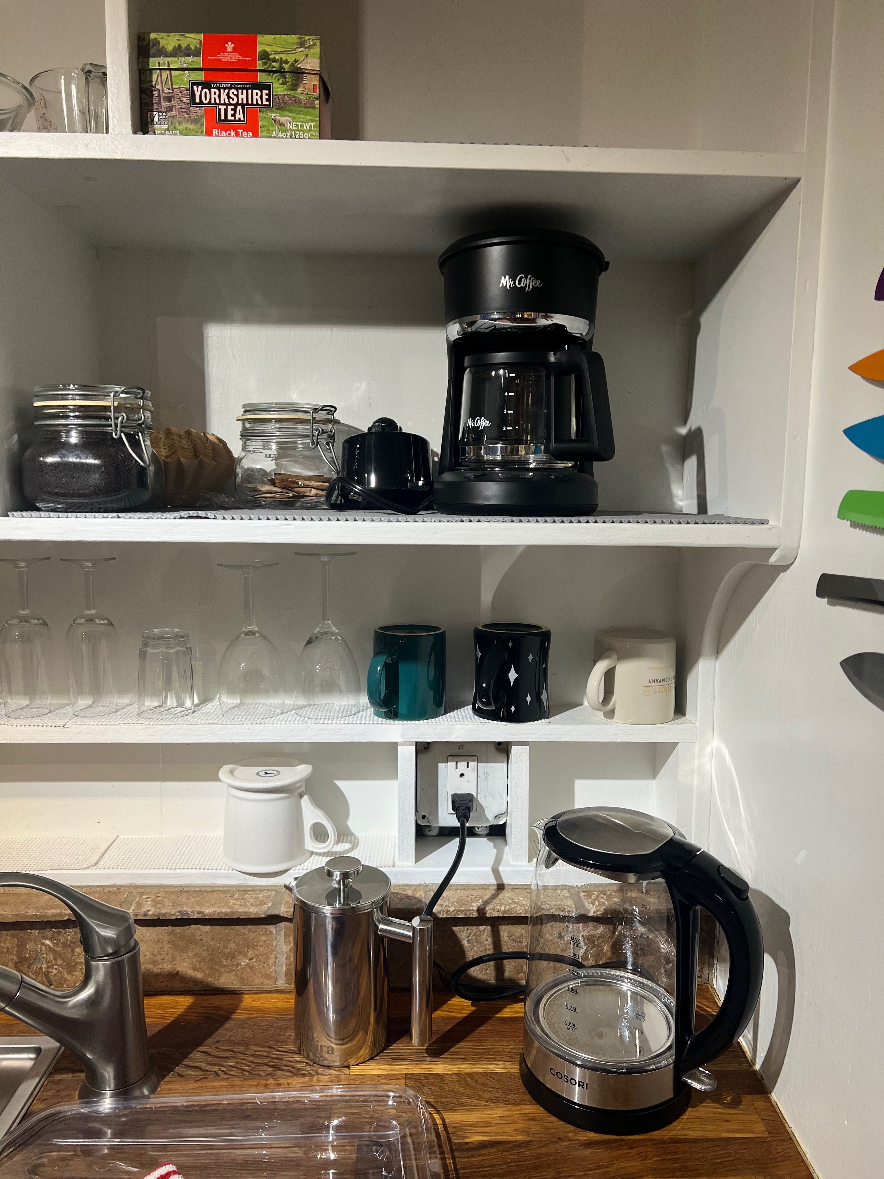 A kitchen area with a coffee maker, jars, and mugs on shelves.