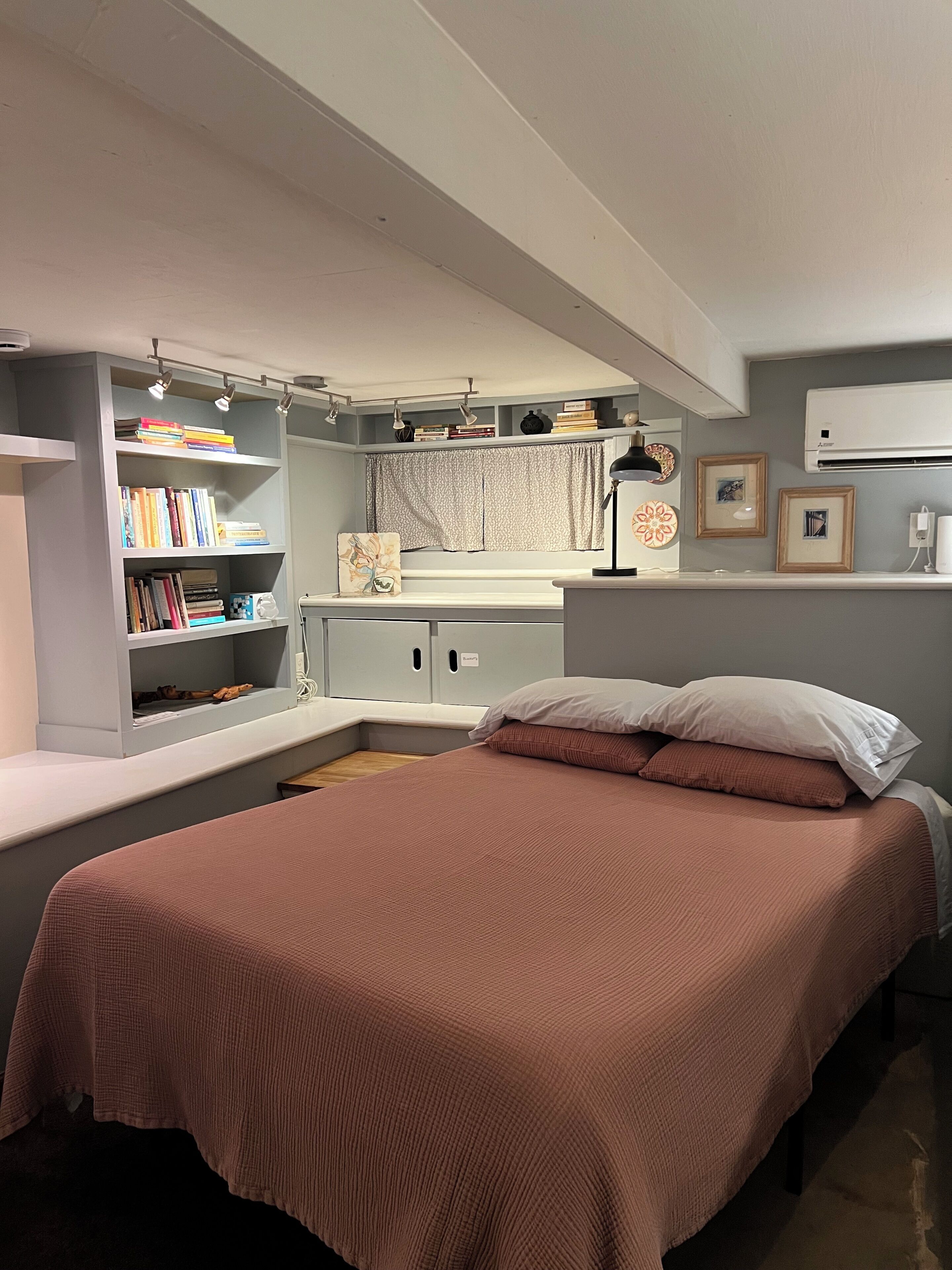 A bedroom with a bed, a bookshelf, a desk, and an air conditioning unit.