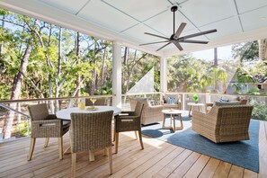 Un patio techado al aire libre con mobiliario de mimbre, un ventilador de techo y vistas a los árboles circundantes.