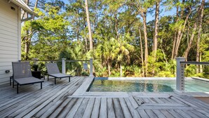 Eine Holzterrasse mit zwei Liegestühlen, einem Swimmingpool und Blick auf hohe Bäume.