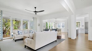 Ein geräumiges Wohnzimmer mit einer großen weißen Couch, einem Deckenventilator und Blick ins Freie durch große Fenster.