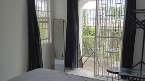 A bedroom with a large bed, a chair, a table, and a view of the outside through a window with curtains. - Charming One-Bedroom Apartment in Peaceful Telescope, St. Andrews, Grenada (Telescope)