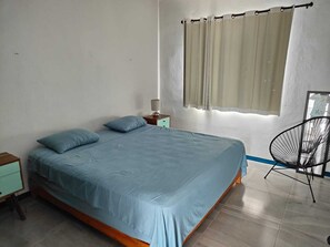 Ein Schlafzimmer mit Bett, Nachttisch, Sessel und einem Fenster mit Vorhängen.