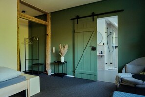 A compact room with a bed, a chair, a small table, a hanging clothes rack, a wall-mounted shelf, and a door leading to a shower area. - House by the lake with plenty of space, right next to the Lüneburg Heath parks (Rethem)