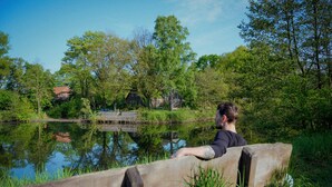 A person sitting on a wooden bench by a calm lake with trees and houses in the background. - House by the lake with plenty of space, right next to the Lüneburg Heath parks (Rethem)