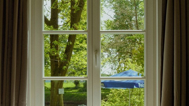 A bedroom with a large window, a bed with pillows, and a view of a park with trees and a blue umbrella. - House by the lake with plenty of space, right next to the Lüneburg Heath parks (Rethem)