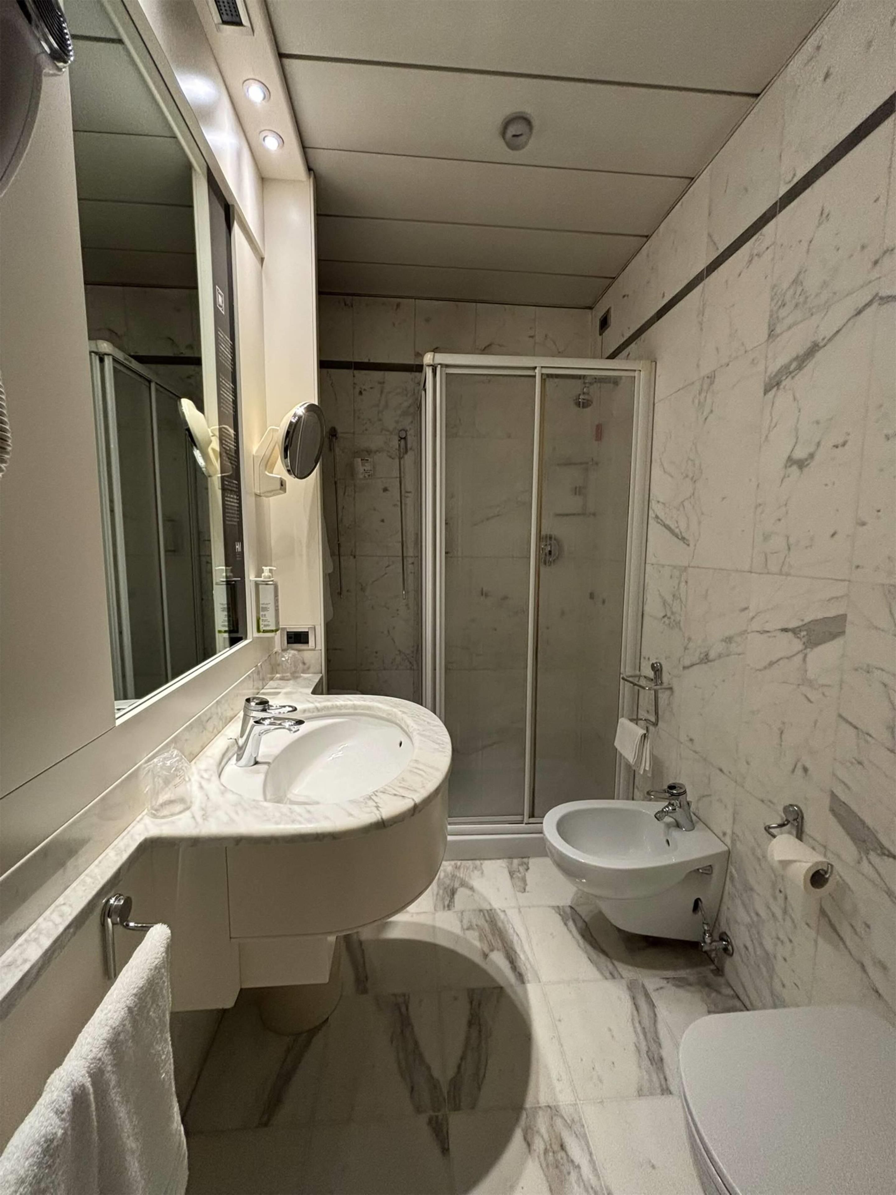A modern bathroom with a large mirror, a marble sink, a bidet, and a shower enclosure.