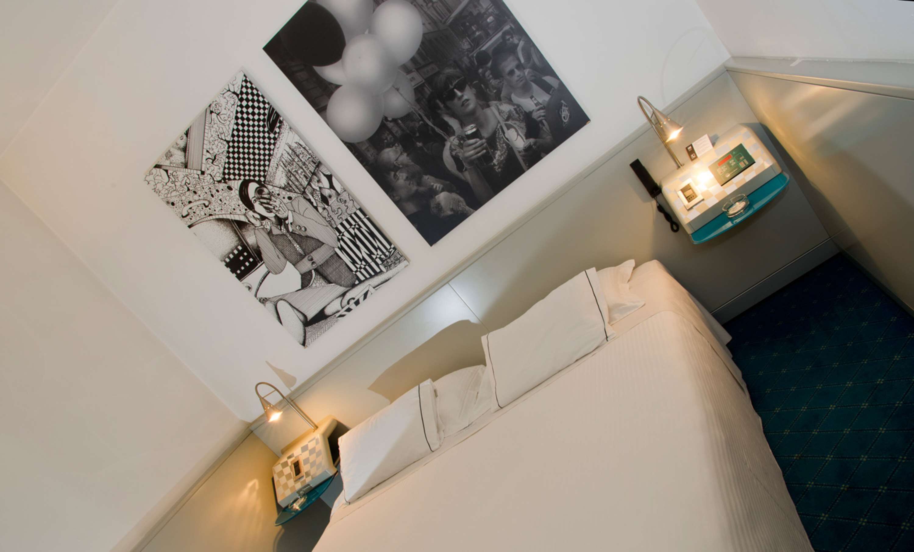 Una habitación de hotel con cama, una mesita de noche con lámpara, un teléfono en la pared y dos cuadros enmarcados.