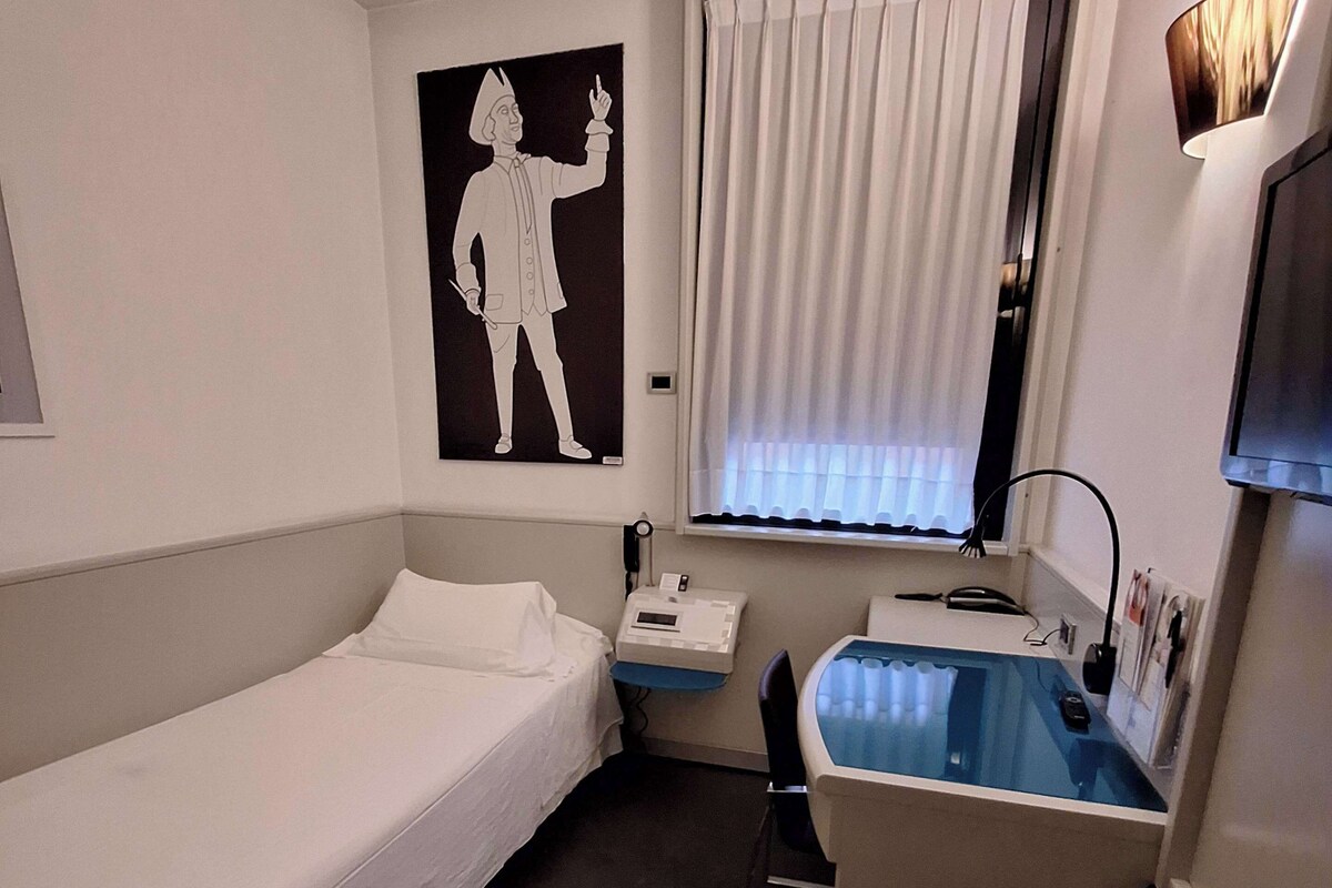 A hotel room with a single bed, a desk with a chair, a window with curtains, a flat-screen TV mounted on the wall, and a poster on the wall.