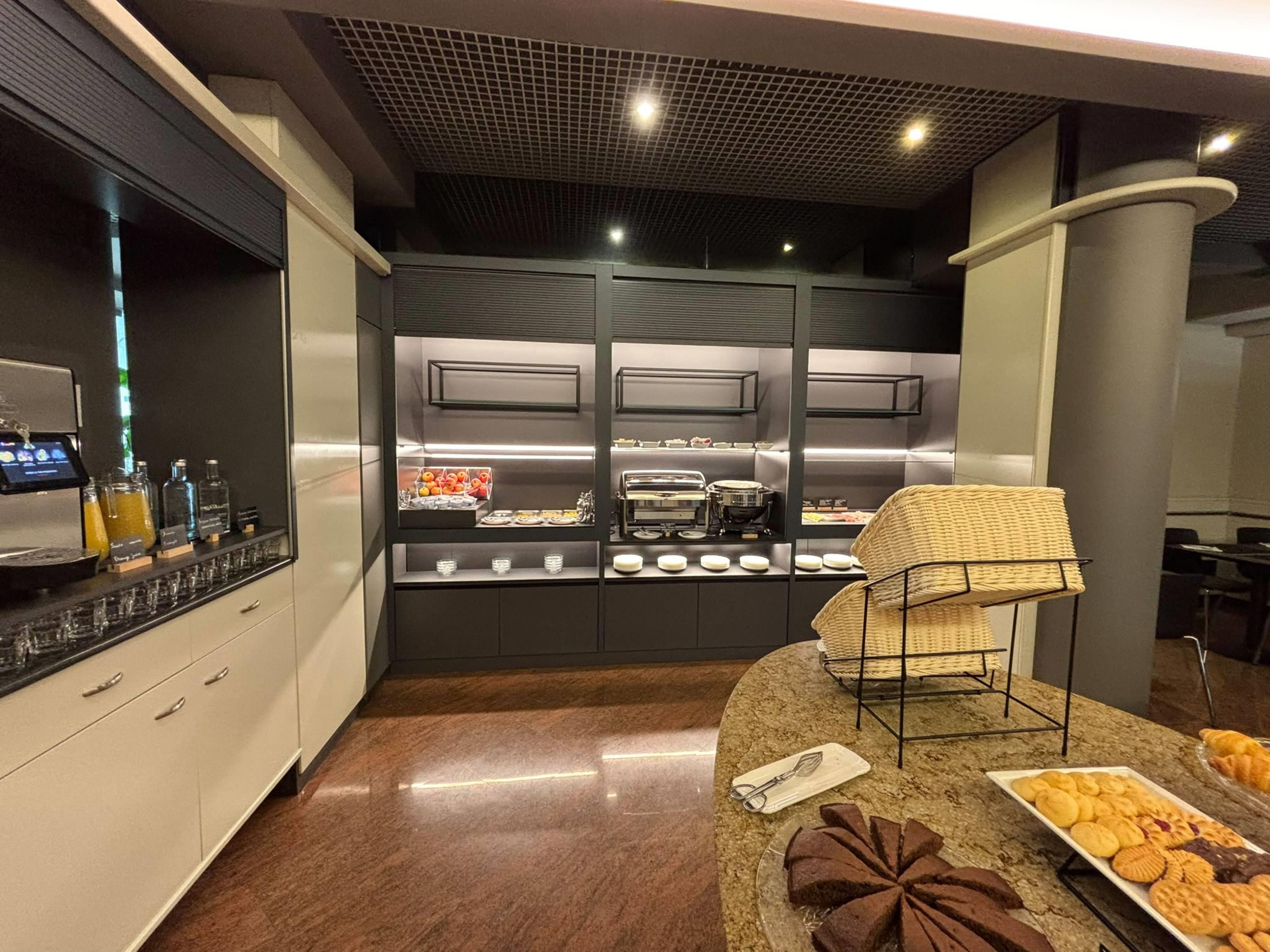 A modern kitchen with a built-in coffee station, a buffet area with various food items, and a seating area with a woven chair.