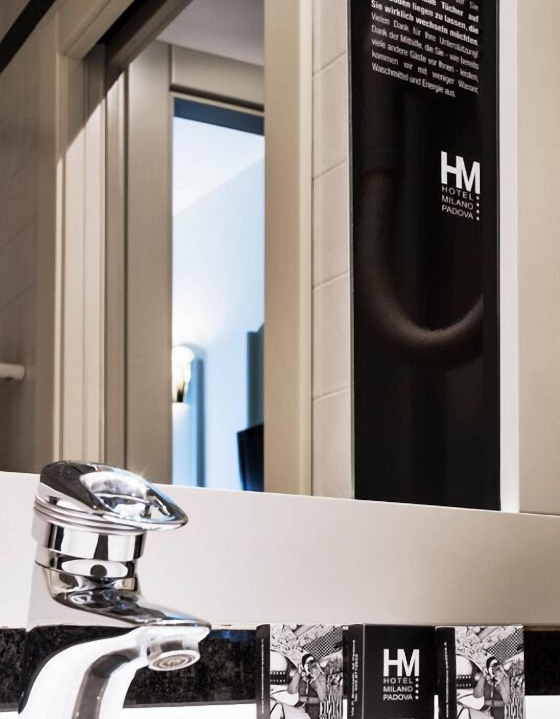 Een moderne badkamer met een chromen kraan, een zwart verticaal bord met de tekst 'HM HOTEL MILANO PADOVA', en een spiegel die de ruimte weerspiegelt.