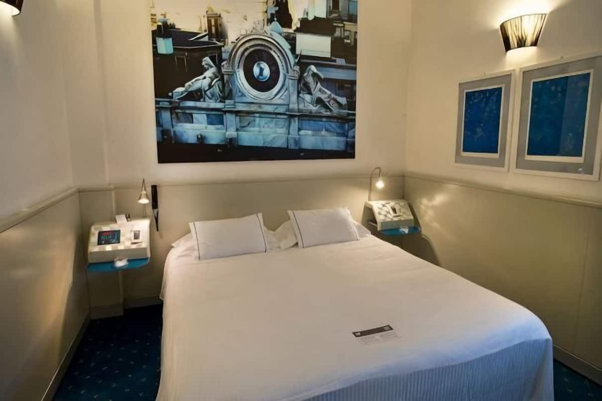 Una habitación de hotel con una cama grande, mesitas de noche, un reloj en la pared y cuadros enmarcados.