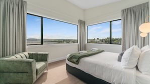 Quarto de hotel moderno com uma cama grande, uma poltrona verde e vista para o horizonte urbano através de amplas janelas.