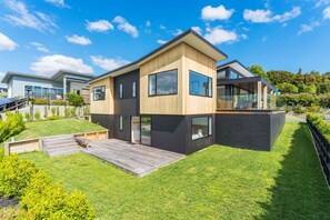 Une maison moderne à deux étages, dotée d’une terrasse en bois, de grandes fenêtres et d’une pelouse bien entretenue.