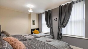 A hotel room with two beds, a headboard, a small bedside table, a clock, and a window with curtains. - City Centre 2bed Apartment Wi-fi Parking (Newcastle-upon-Tyne)