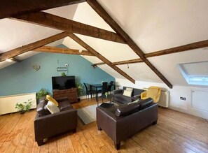 Een loft-woonkamer met zichtbare houten balken, een flatscreen televisie, donkere banken en een eettafel met stoelen.