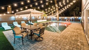 Un patio trasero con piscina, mobiliario exterior e iluminación con luces colgantes.