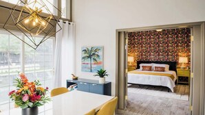 Una habitación de hotel moderna con una cama grande, un área de comedor con una mesa y sillas, y vistas a través de una puerta abierta a un dormitorio con una pared colorida.