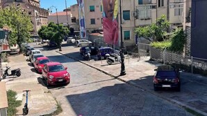 Vista de una calle con coches y scooters estacionados, edificios con balcones y cielo despejado.