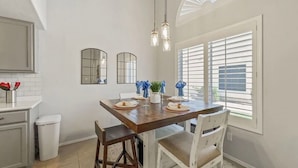 A dining area with a wooden table, white chairs, and a chandelier. There are two wall-mounted mirrors and a window with blinds. - Perfect Chandler Getaway – Cozy Condo in the Heart of the East Valley! (Chandler)