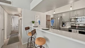 A modern kitchen with white countertops, stainless steel appliances, and two bar stools. - Perfect Chandler Getaway – Cozy Condo in the Heart of the East Valley! (Chandler)