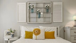 A bedroom with a white bed, yellow pillows, a large yellow flower decoration, and a window with white shutters displaying small vases with flowers. - Perfect Chandler Getaway – Cozy Condo in the Heart of the East Valley! (Chandler)