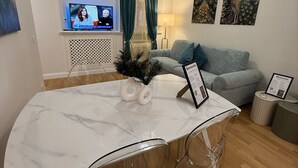 Ein modernes Wohnzimmer mit einem Marmorestisch, klaren Stühlen, einer Chandelierleuchte, einem Fernseher, einer Couch und dekorativen Pfauenbildern, sowie einem Willkommensschild.