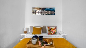 A neatly arranged hotel room with a bed, a bedside table with a lamp, a tray with breakfast items, and a cityscape painting on the wall.