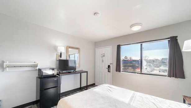 A hotel room with a bed, a desk with a television, a refrigerator, a window with a view of the outside, and a door. - Extended Stay by Welcome INN (Blue Springs)