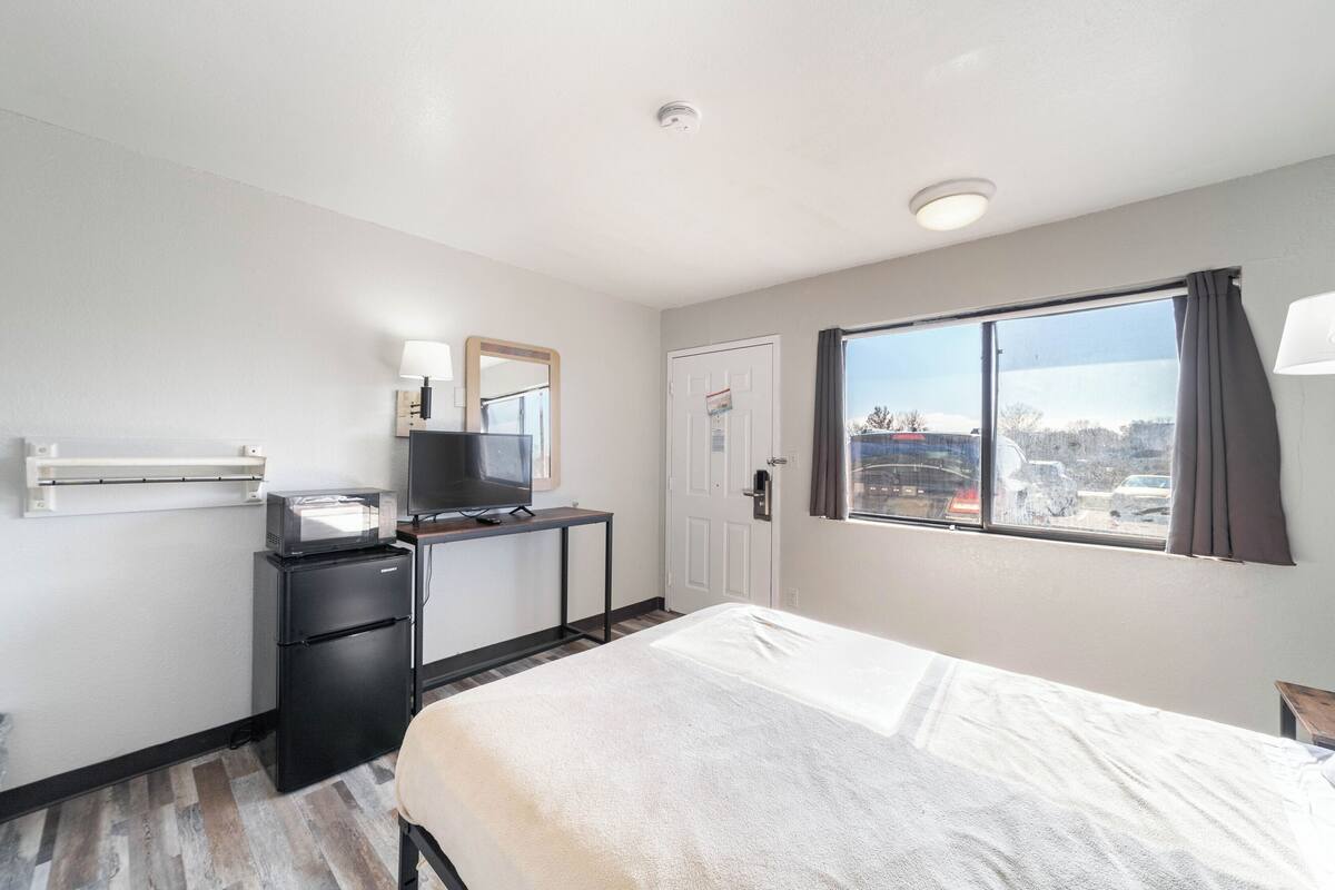 A hotel room with a bed, a desk with a television, a refrigerator, a window with a view of the outside, and a door.