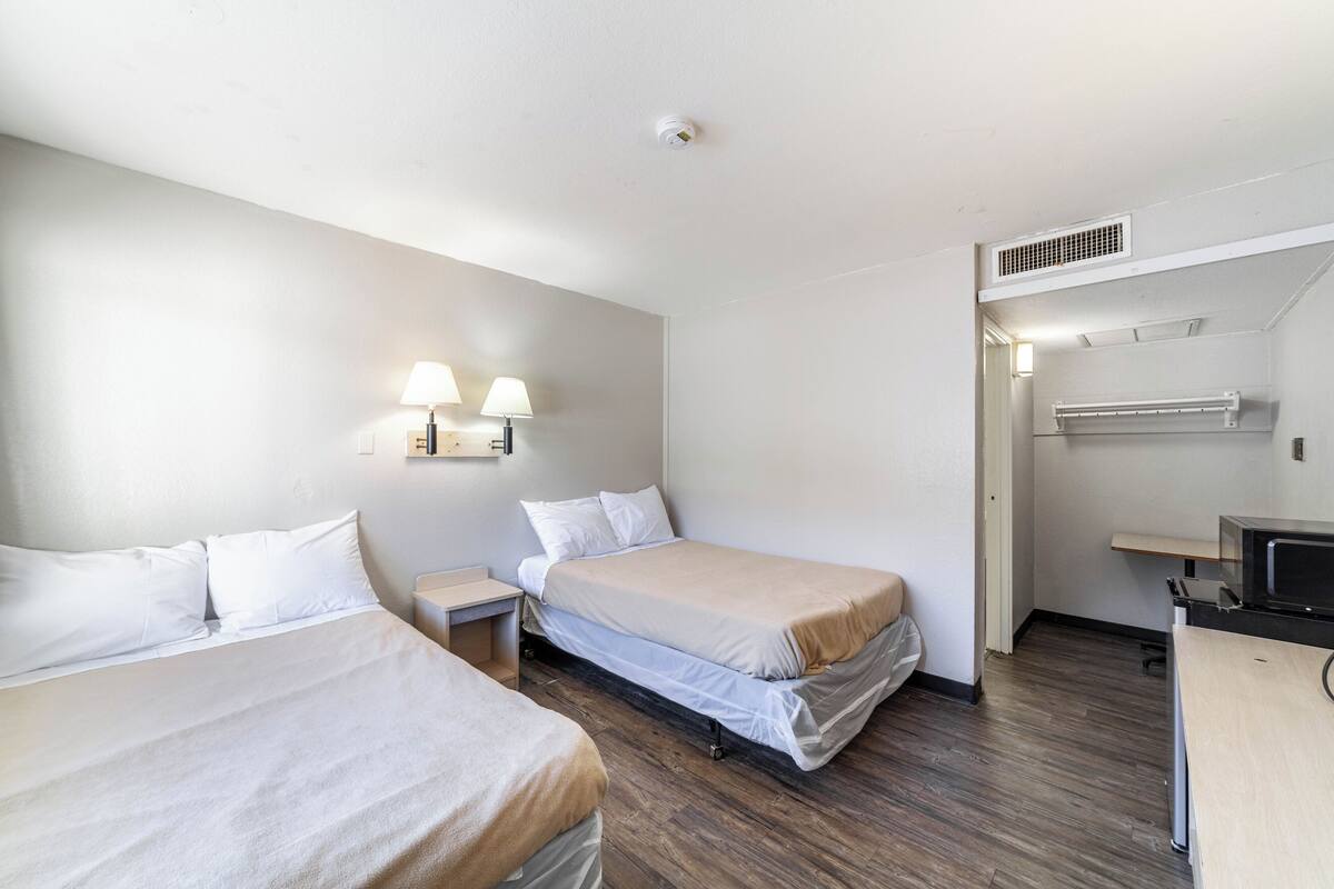 A hotel room with two beds, a desk, a chair, a lamp, a microwave, and a vent.