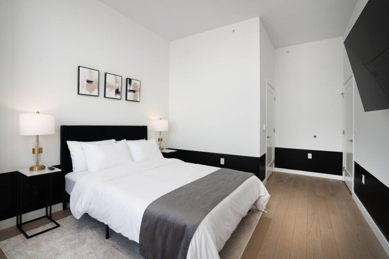 A modern hotel room with a large bed, two bedside tables with lamps, a wardrobe, and a door leading to another room.