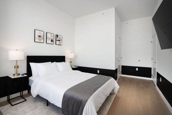 Una habitación de hotel moderna con una cama grande, dos mesitas de noche con lámparas, un armario y una puerta que da a otra habitación.