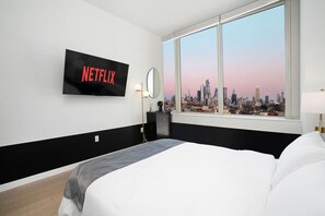 A hotel room with a large bed, a TV mounted on the wall displaying 'NETFLIX', a cityscape view through the window, and a bedside table with a lamp.