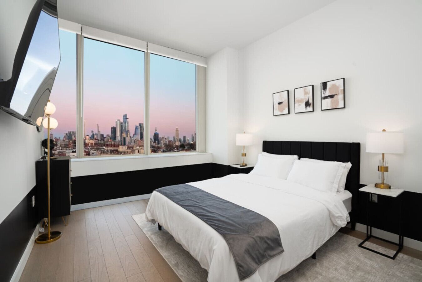 Habitación de hotel con cama, dos mesitas de noche, televisor montado en la pared y un ventanal con vistas a la ciudad.