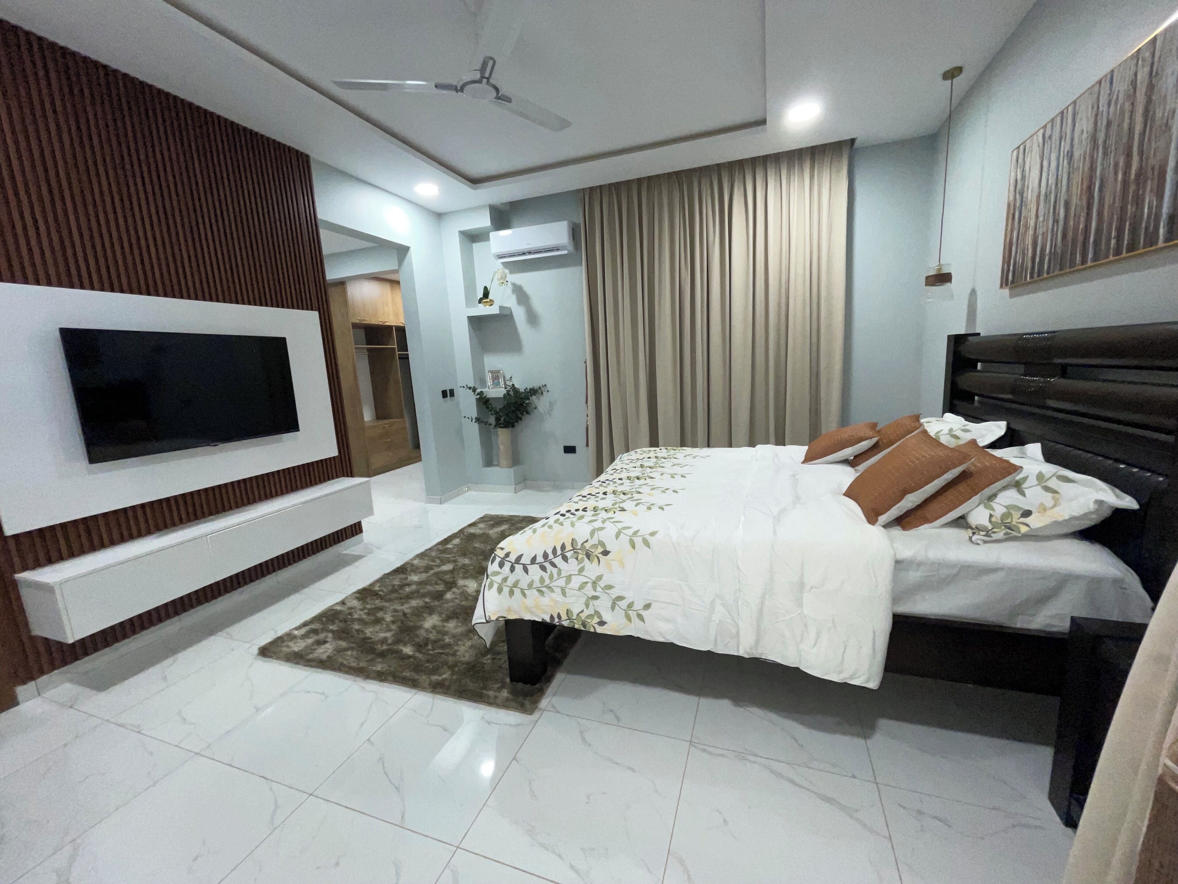 Une chambre à coucher moderne avec un téléviseur à écran plat fixé au mur, un lit recouvert d’une couvre-lit à motifs, une tête de lit sombre et un ventilateur de plafond.