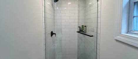 A narrow shower area with white tiled walls, a black showerhead, and a shelf with toiletries.