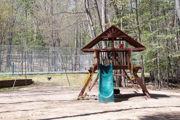 Ein Spielplatz mit einer Holzspielkonstruktion, darunter eine Rutsche und Schaukeln, umgeben von Bäumen.