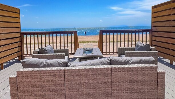 A wooden deck with a wicker sofa and a table, overlooking a beach. - NEW! The Shipwreck Studio (Corpus Christi)