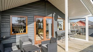 A wooden cabin with a covered patio area featuring wicker furniture and a glass door leading to a deck.
