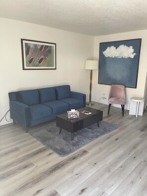 Un salón con un sofá azul, una mesa de centro con una caja de pañuelos, una lámpara y un cuadro de nubes.