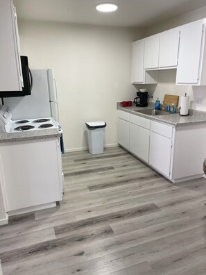 Una cocina con armarios blancos, una estufa, un bote de basura y una encimera con cafetera y jabón para lavar platos.