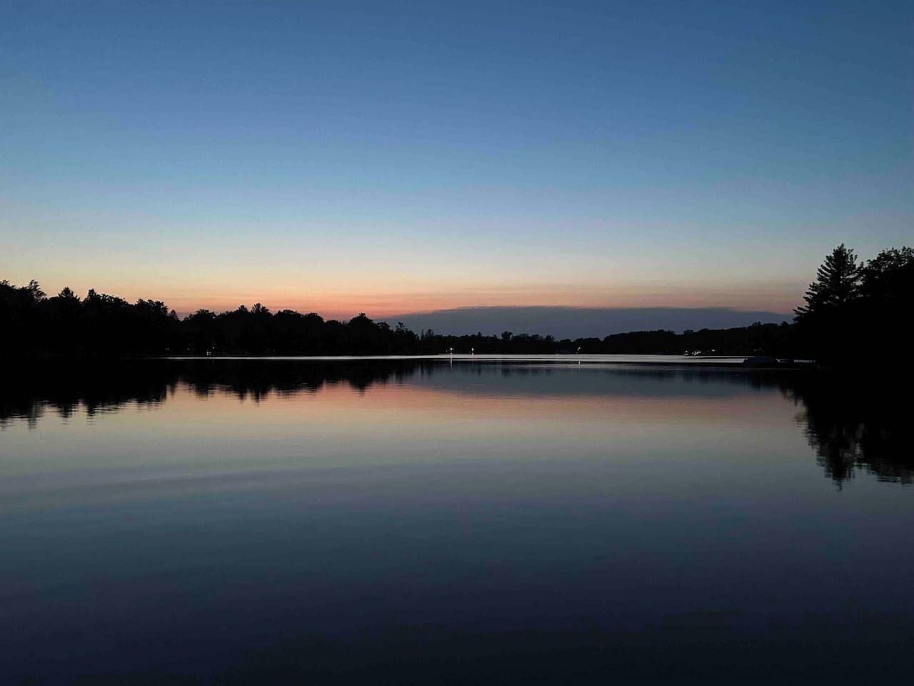 A calm lake at dusk with a clear sky and distant trees.