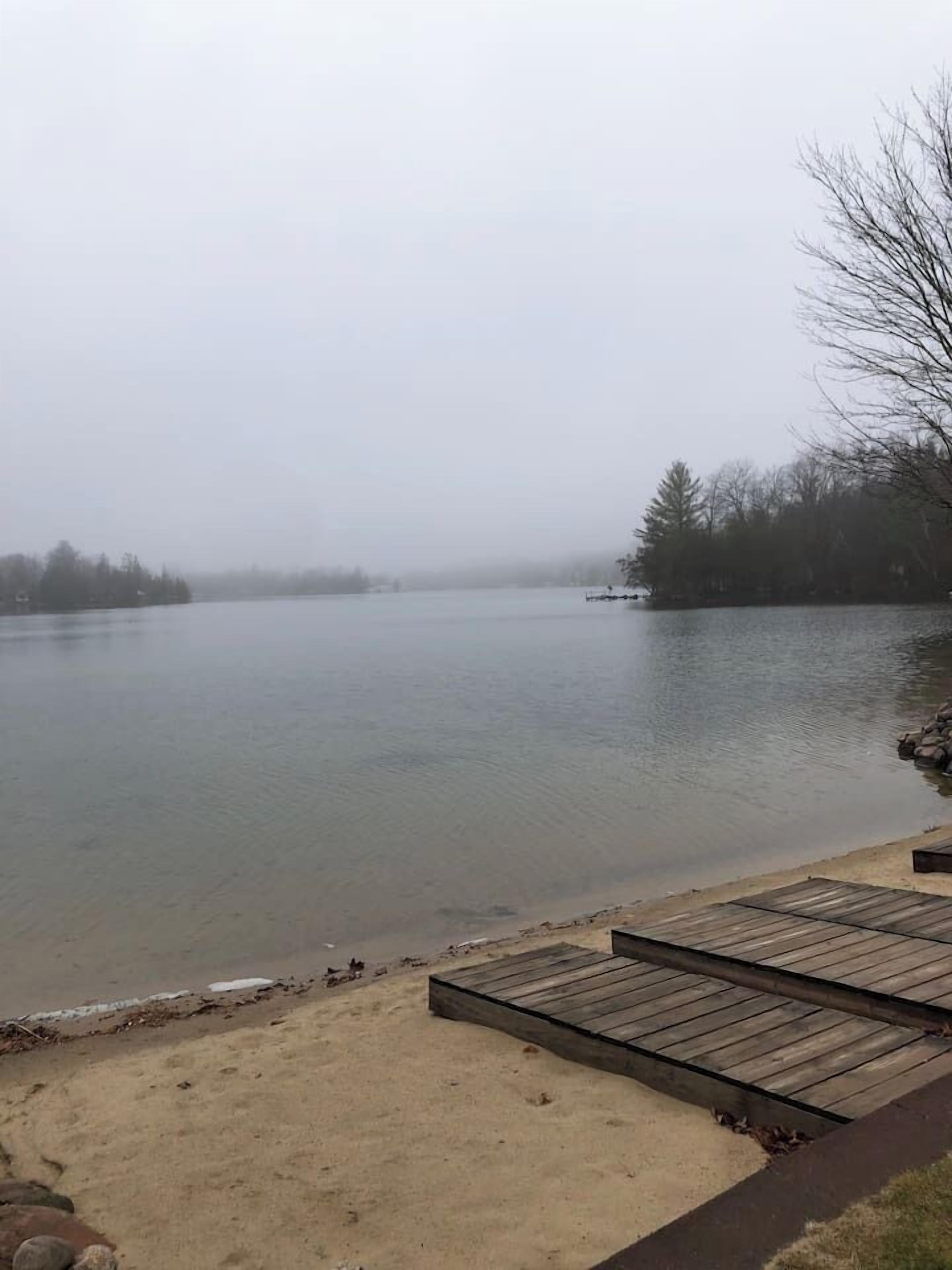 Un lac avec une jetée en bois et une plage de sable, entouré d’arbres et surplombé d’un ciel nuageux.