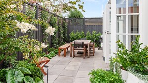 Een terras met een houten tafel en stoelen, omringd door weelderige begroeiing en bloeiende planten.