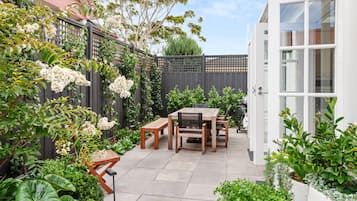 A patio with a wooden table and chairs, surrounded by lush greenery and flowering plants.