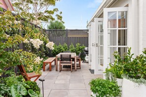 Un patio con mesa y sillas de madera, rodeado de abundante follaje y plantas en flor.
