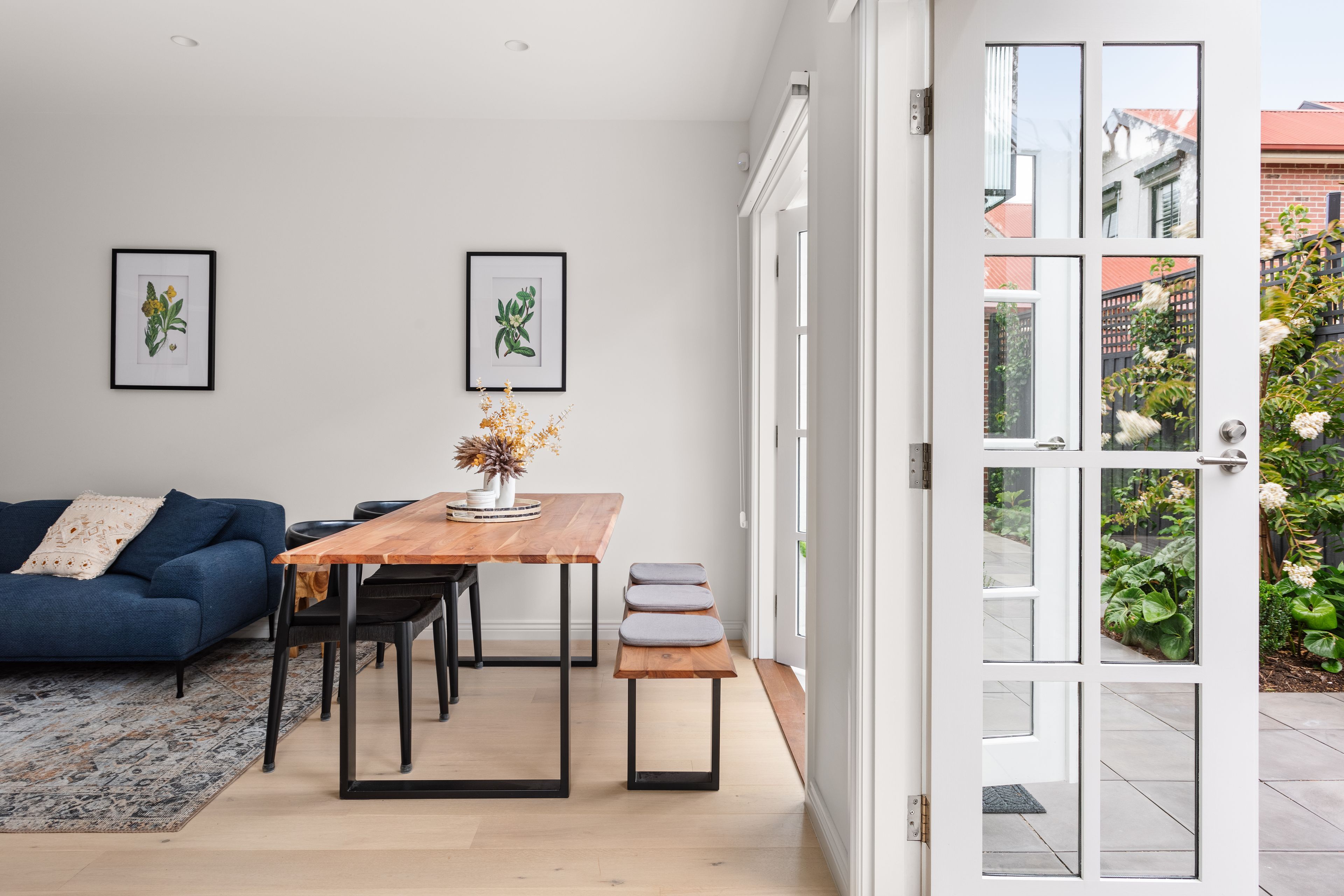 Un salon moderne avec une table à manger en bois, un canapé bleu et deux reproductions botaniques encadrées accrochées au mur.