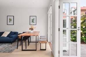 A modern living room with a wooden dining table, a blue sofa, and two framed botanical prints on the wall.